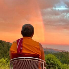 Jigme Rinpoche seated facing a sunset rainbow over the ocean